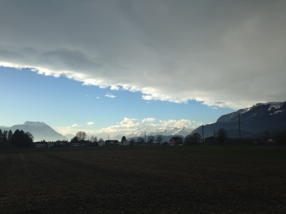 Blick über ein Feld mit Bergen auf der Seite und im Hintergrund. Der Himmel ist zur Hälfte blau, die andere Hälfte ist mit einer grauen Wolkenschicht bedeckt.