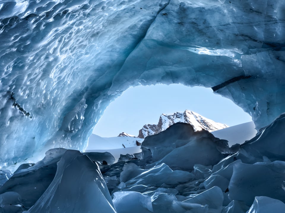 Eishöhle aus blauem Eis. In der Bildmitte der Ausgang der Höhle, draussen eine winterliche Berglandschaft.