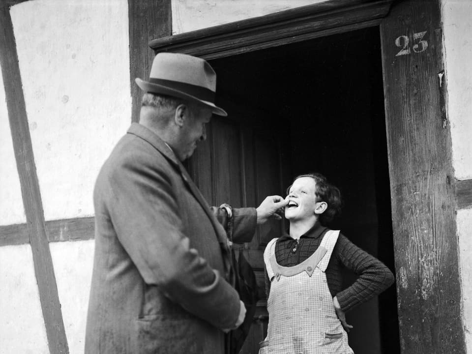 Schwarzweiss Aufnahme: Mann begutachtet die Zähne eines Mädchens, das vor einem Hauseingang steht.