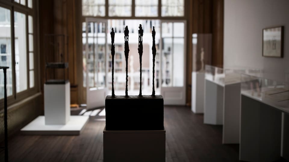Vier dünne Skulpturen im Gegenlicht in einem Museumsraum mit Virtrinen und Tafeln.