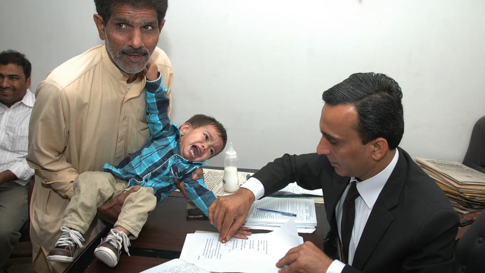 Ein Mann hält ein Kleinkind, während ein anderer Mann ein Papier mit dessen Fingerabdrücken versieht.