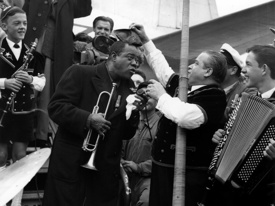 Louis Armstrong, der aus dem Flugzeug aussteigt und von Schweizer Volksmusikern empfangen wird
