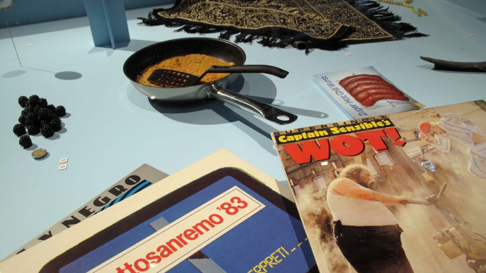 Stilleben mit einer Pfanne mit Rösti drin, einem Reisekatalog und einer Zeitschrift.