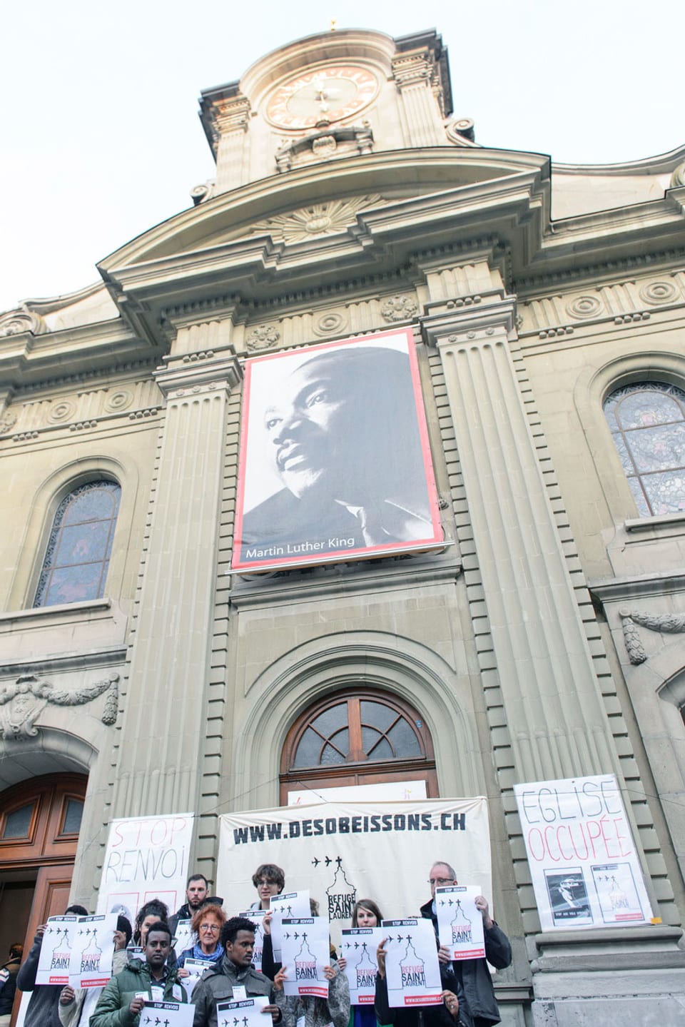 Menschengruppe mit Plakaten steht vor Kirche.