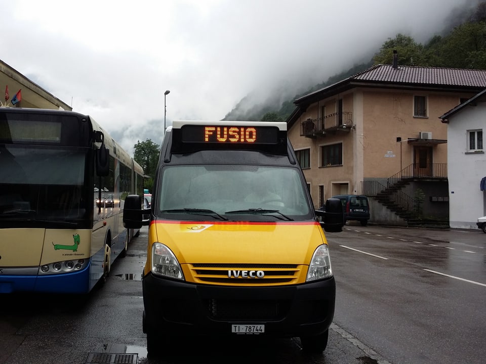 Ein Kleinbus in den Farben des Postautos steht am Bahnhof bei Regenwetter.