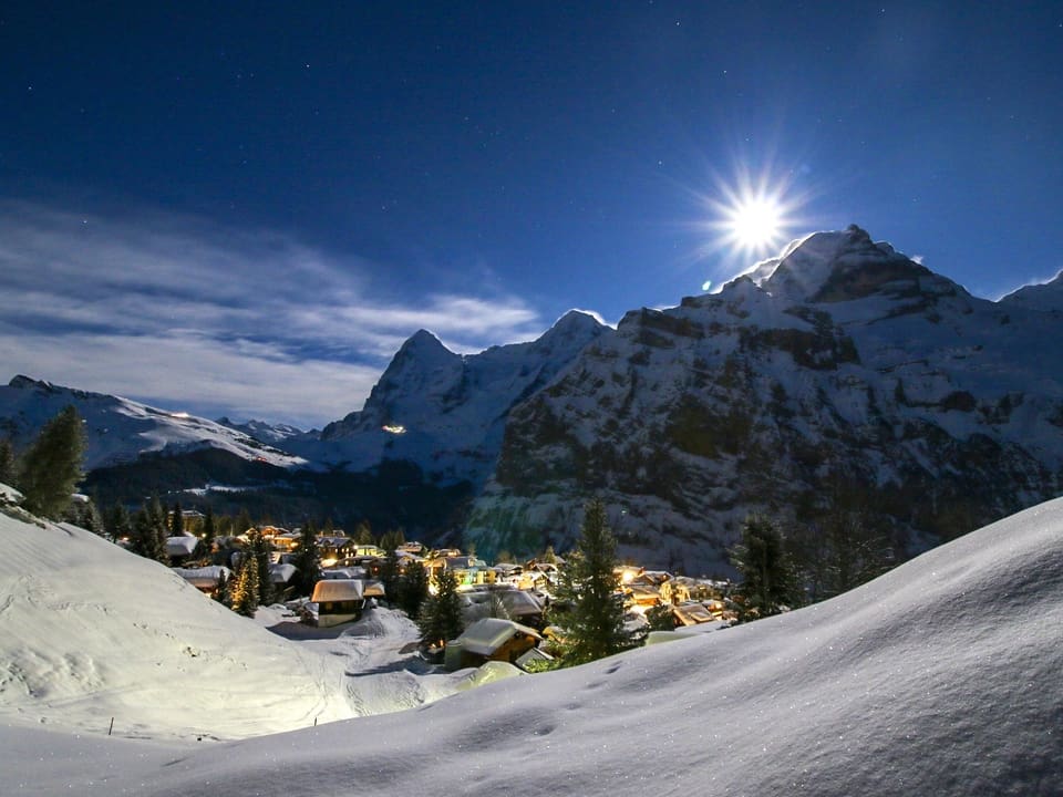 Nacht in den Alpne mit viel Schnee. Der Mond leuchtet und auch die Lichter eines Dorfes sind zu sehen. 