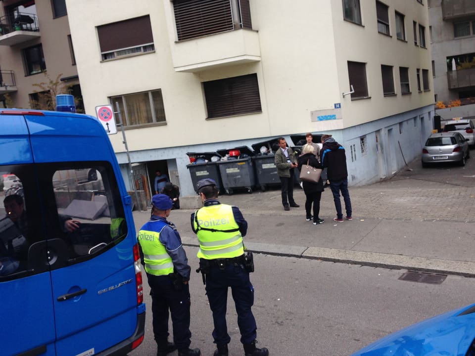 Zwei Polizsten neben ihrem blauen Kastenwagen vor einem Wohnhaus, dort stehen weitere Personen vor überfüllten Abfallcontainern.