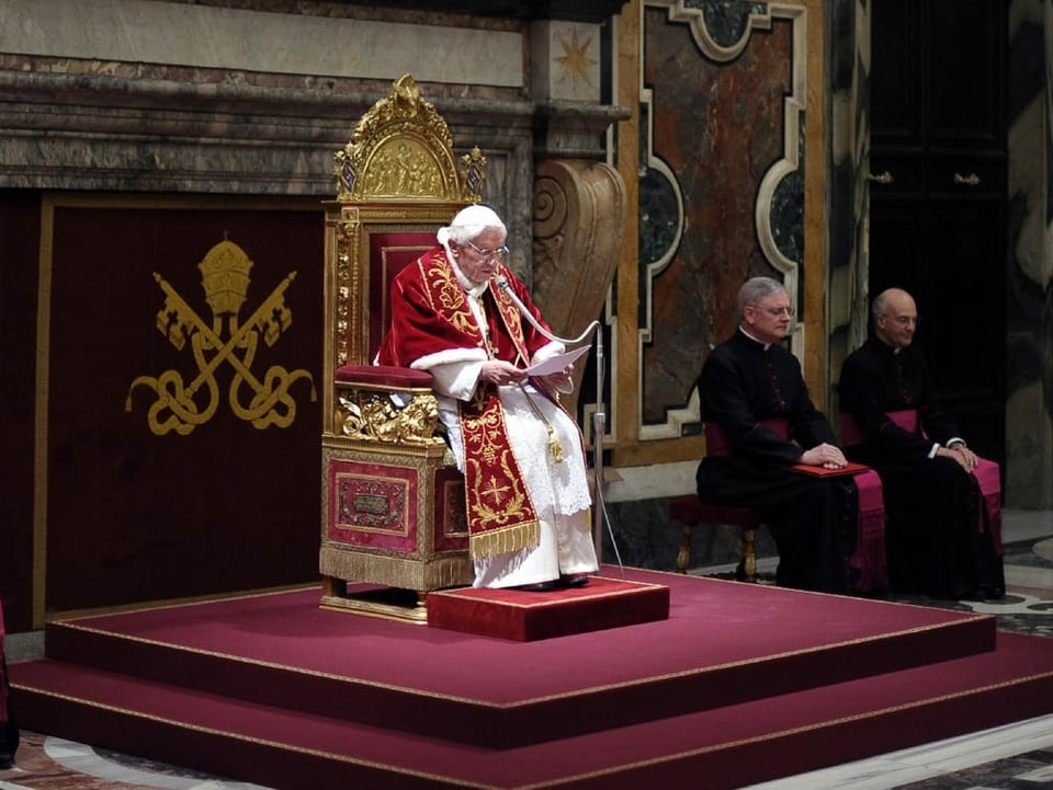 Der Papst sitzt auf einem geschmückten Stuhl und liest von einem Papier ab.