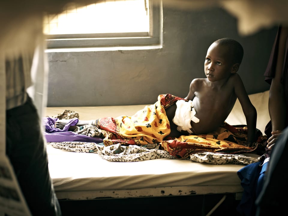 Ein Kind liegt in einem Spital in Tansania auf einem Bett, Tücher um sich.