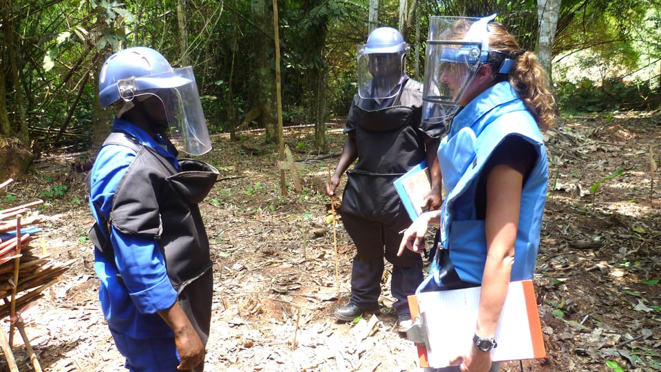 Drei Kampfminenbeseitiger stehen mit Schutzkleidung in einem Kreis im Wald und besprechen sich