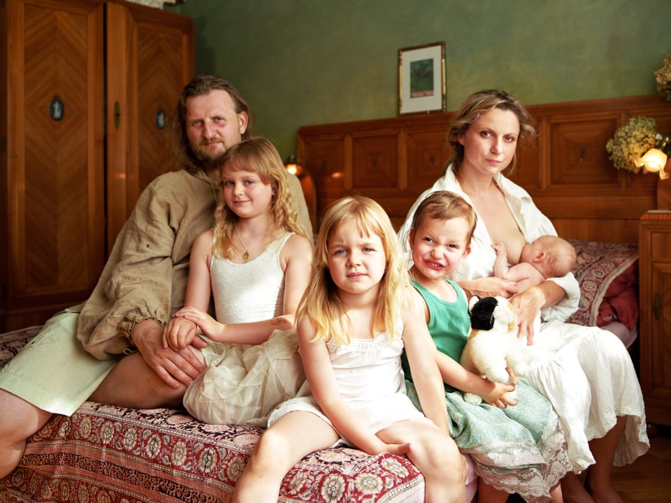 Ein Mann und eine Frau sitzen mit vier Kindern auf einem Bett, das jüngste wird von der Frau gestillt.