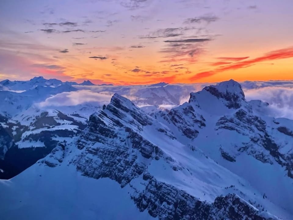 Blick über verschneite Alpen mit gelb bis violettem Himmel. 