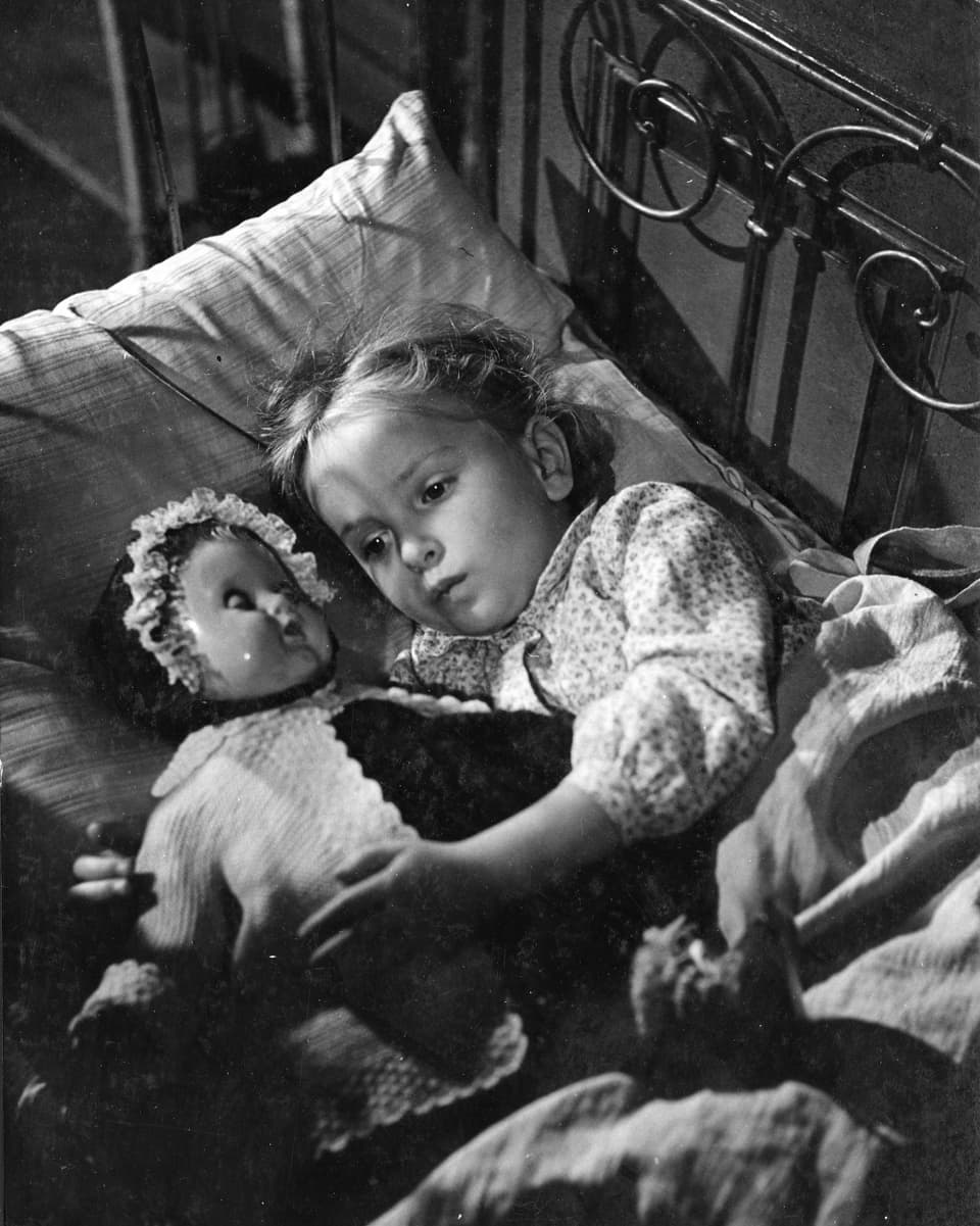 Ein junges Mädchen liegt mit ihrer Puppe im Bett, ihre Augen sind geöffnet.