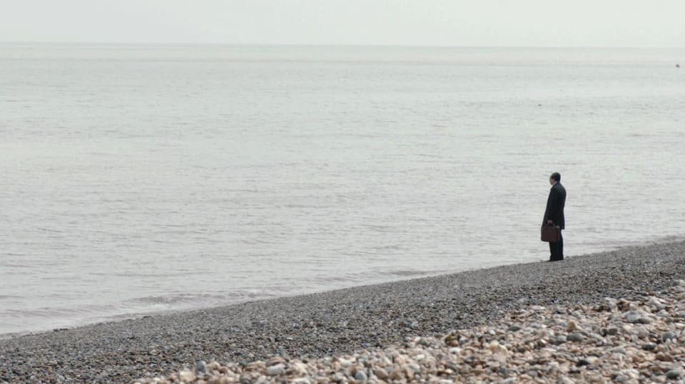 Ein Mann steht an einem Kieselstrand und blickt ins graue Meer hinaus.