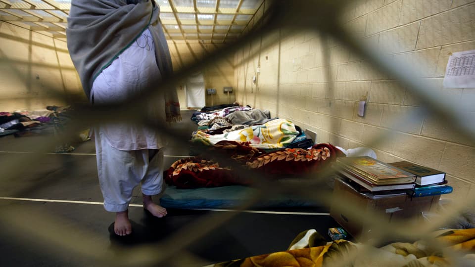 Mann in einer Zelle für mehrere Häftlinge mit mehreren Matratzen am Boden. Decken liegen herum.