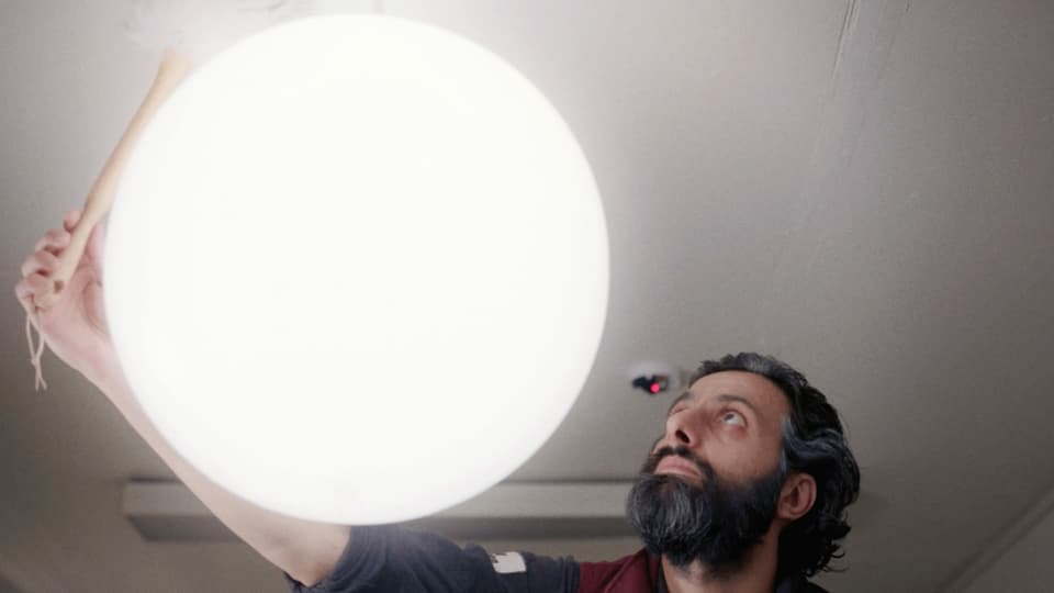 Ein Mann mit Bart wischt eine grosse, runde Deckenlampe mit einem Staubwedel.