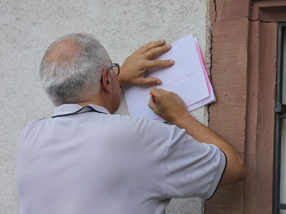 Ruedi Matter hält ein Blatt Papier an eine Hauswand und schreibt darauf.