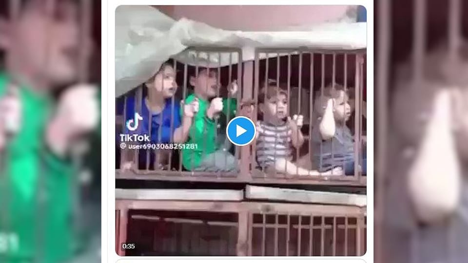 Video-Still von vier kleinen Kindern in einem rostigen Käfig, halten teils die Gitterstäbe, schauen nach draussen.