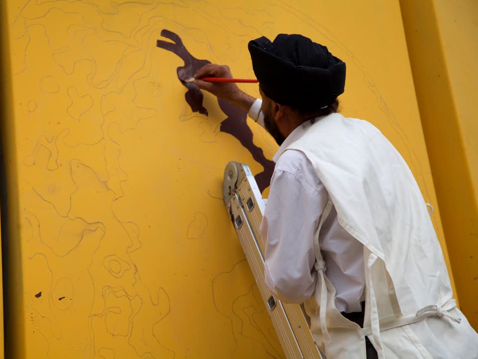Mann auf Leiter an einer gelben Betonmauer malend