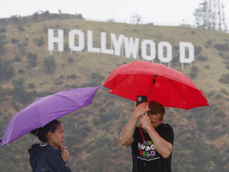 Zwei Personen halten Regenschirme fest; im Hintergrund Hollywood-Schriftzug
