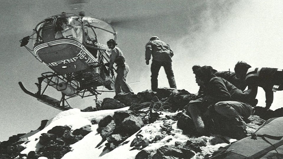 Ein Helikopter rotiert über dem Schreckhorn. Die Alpinisten erwarten in kauernder Stellung die Leute von Radio und Fernsehen.