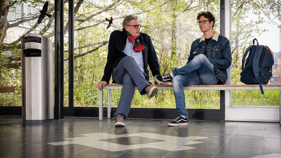 Vater und Sohn sitzen auf einer Bank im Flur eines Schulgebäudes und unterhalten sich.