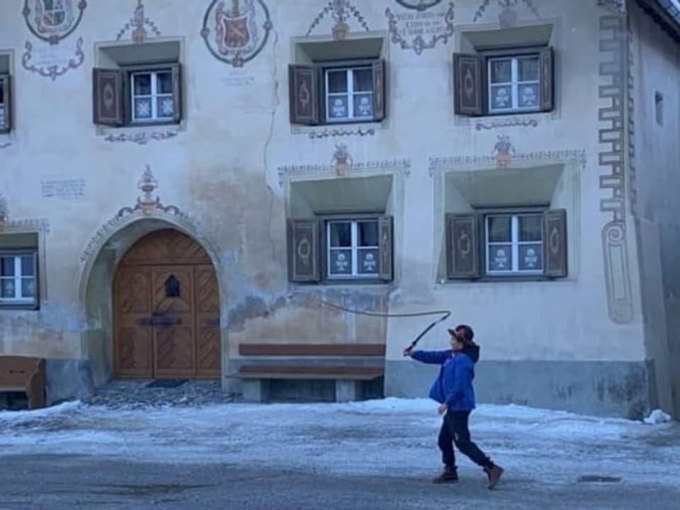 Ein Junge steht vor einem traditionellen engadiner Haus und schwingt seine Peitsche.