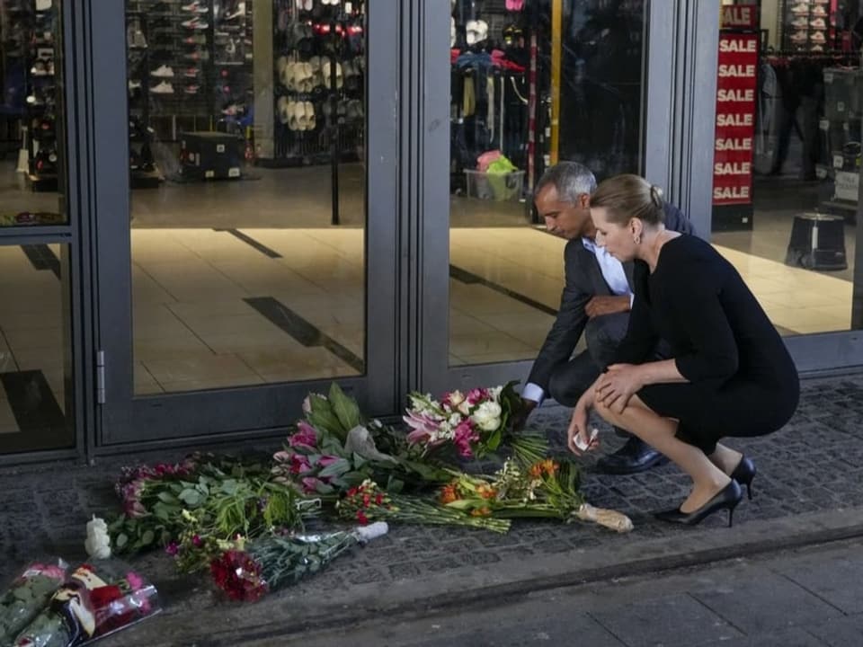 Beide kauern vor dem Eingangsbereich des Einkaufszentrum und legen einen Blumenstrauss neben fünf andere.