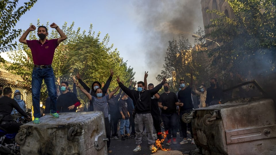Menschen protestieren mit geöffneten Armen und neben brennenden Containern auf den Strassen Teherans.