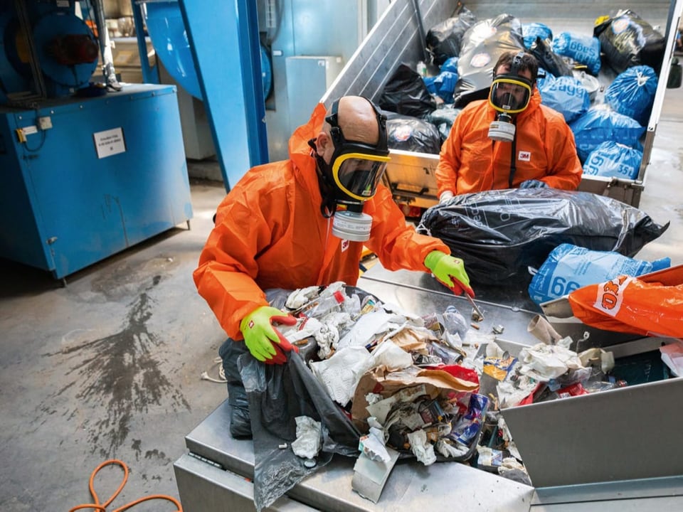 Drei Personen in orangenen Schutzanzügen und Gasmasken durchwühlen Abfall.