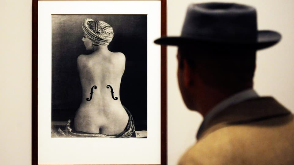Mann mit Hut steht vor dem Foto eines Frauenrückens auf den die Schalllöcher einer Geige gemalt sind. 