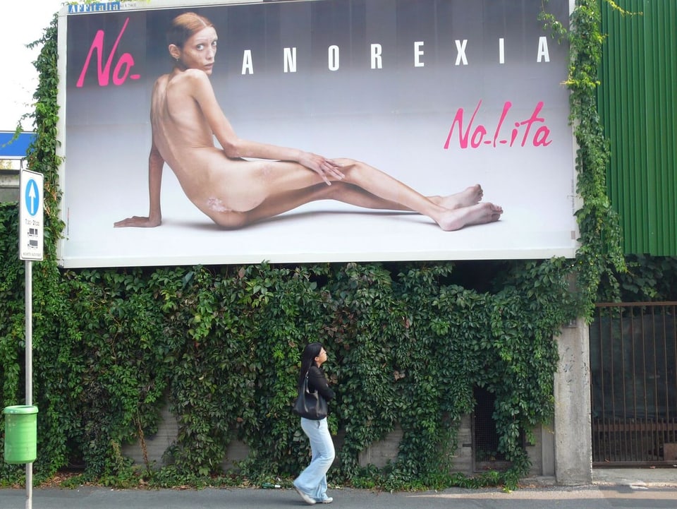 Eine nackte magersüchtige Frau schaut schüchtern von einem Plakat. 