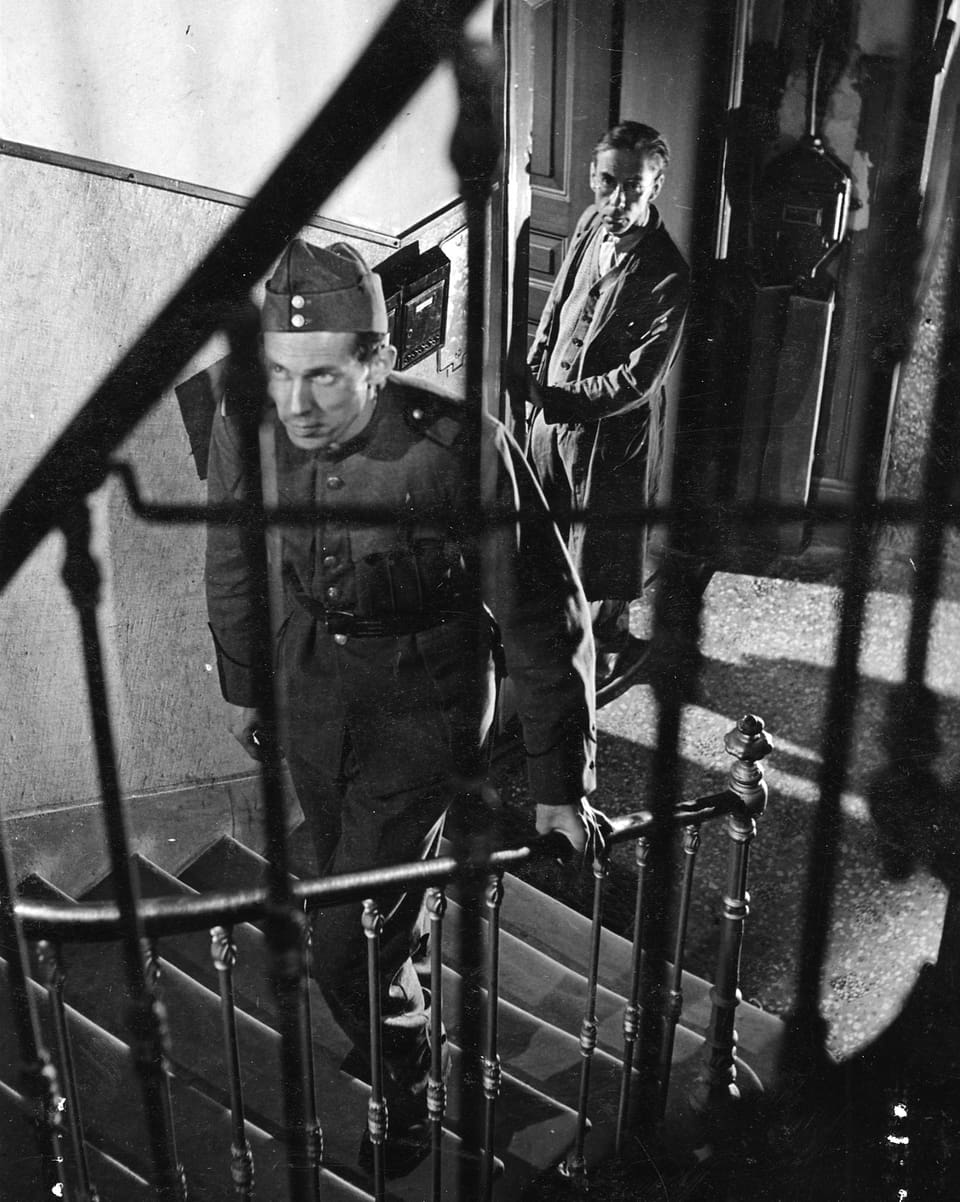 Ein Mann in Militärunform steigt eine Treppe hoch. Ein zweiter Mann beobachtet ihn dabei aus einer Tür im Hintergrund. 