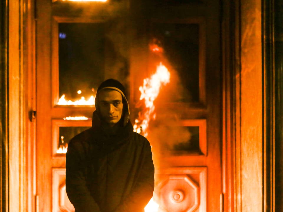 Mann mit Kapuze steht vor brennender Tür.