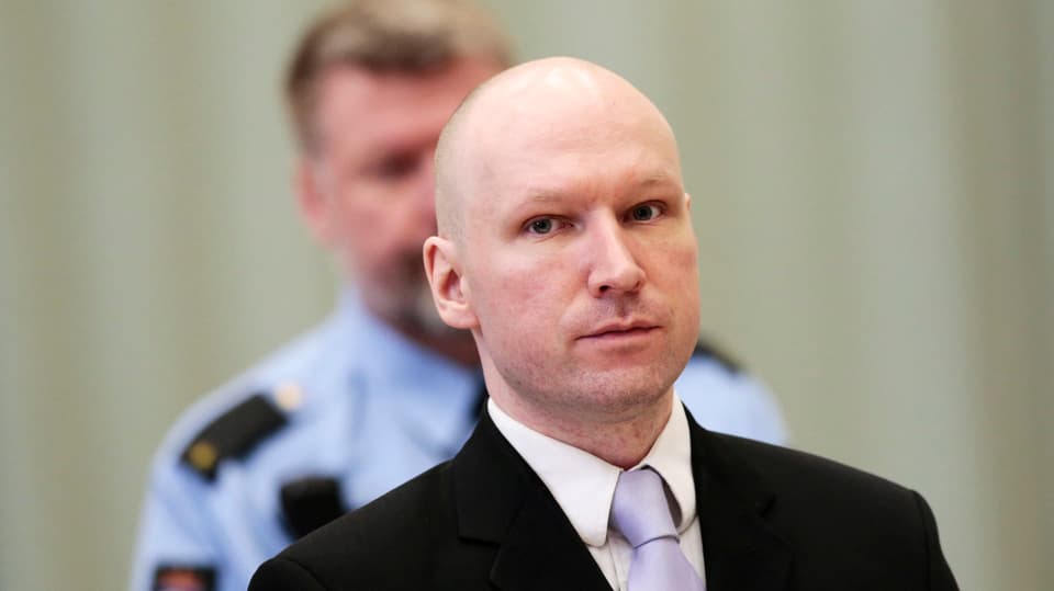 Breivik mit Anzug und Krawatte. Im Hintergrund ein Polizist. 