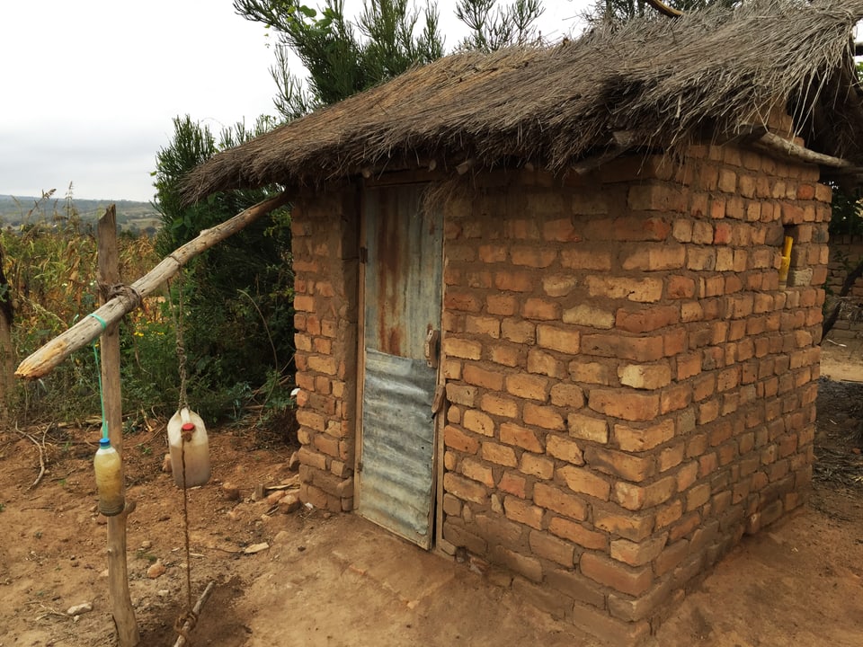 Eine Latrine in Tansania: Eine Hüte aus Bachsteinen ist zu sehen, vor der Tür hängen zwei Flaschen.