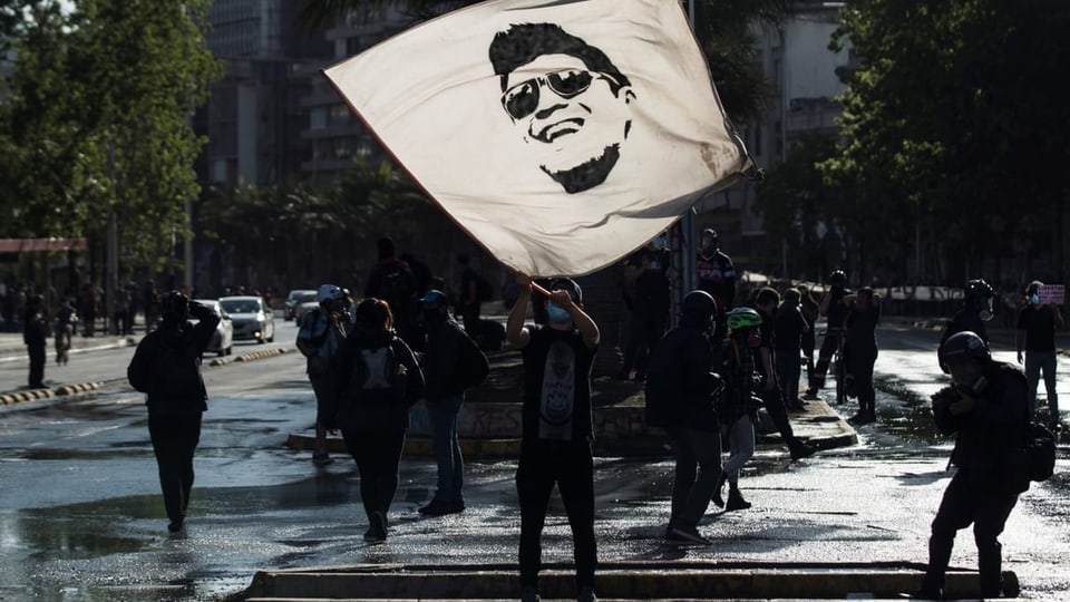 Ein Demonstrant hält eine Flagge in die Luft. Auf dieser ist das Gesicht von Gustavo Gatica zu sehen. 