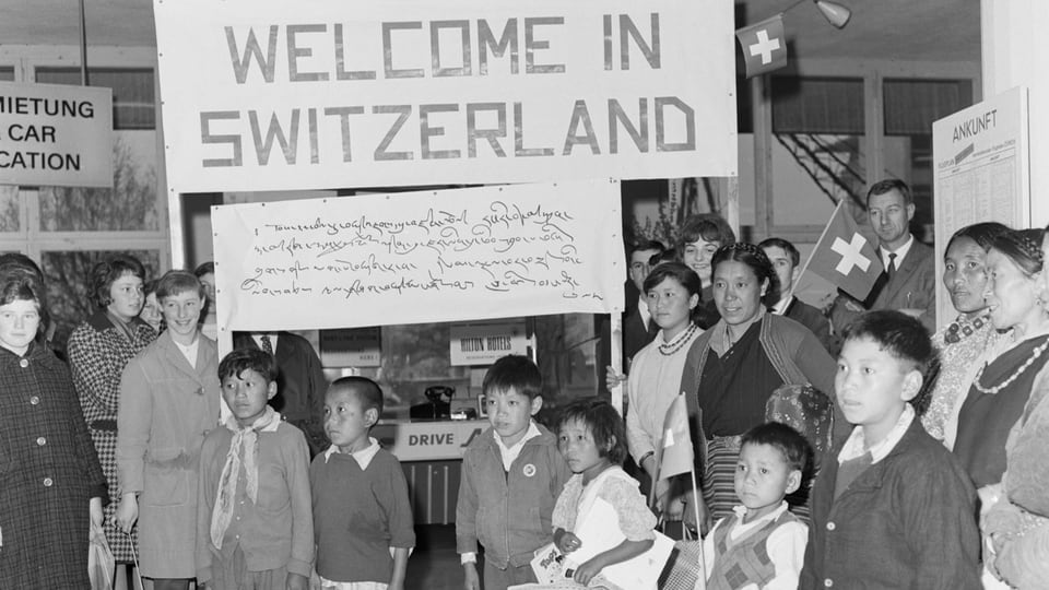 schwarzweiss-Aufnahme mehrere Kinder und hinten Erwachsene stehend, halten Plakat «Welcome to Switzerland»