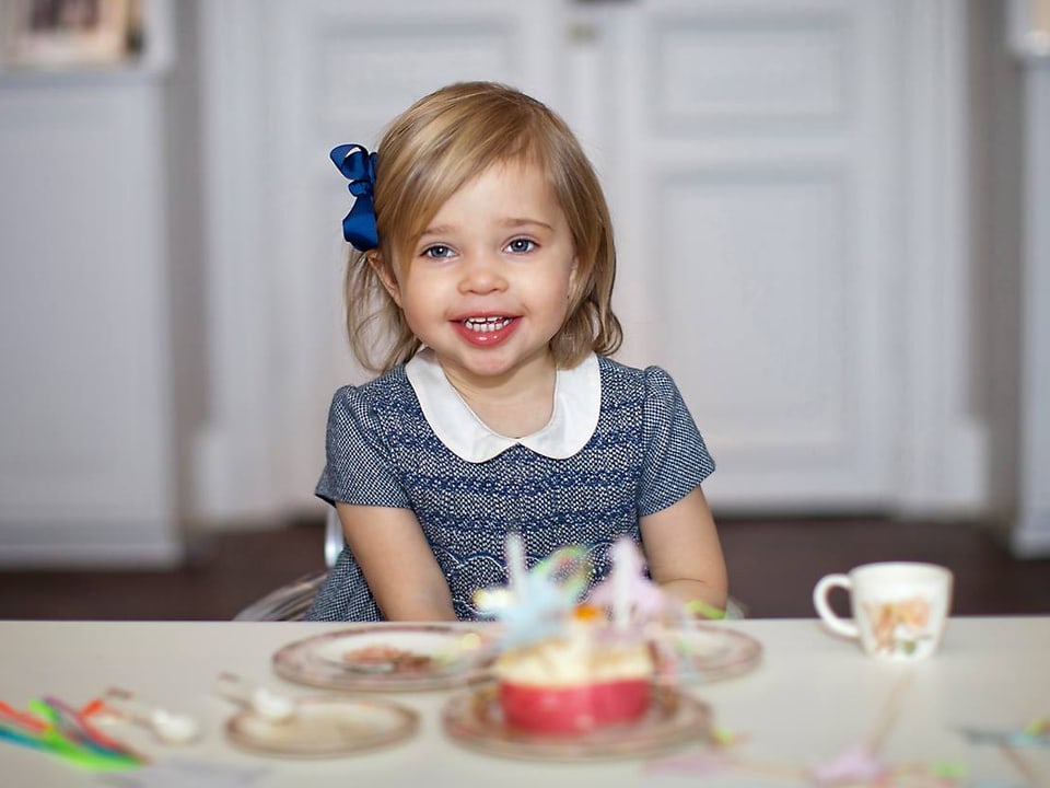 Ein Mädchen in blauem Kleidchen und blonden Haaren lächelt und blick in die Kamera. Sie sitzt an einem gedekten Tisch.