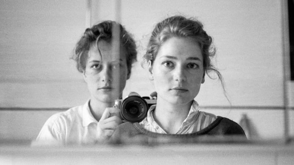 schwarzweiss Foto: Vorne junge Frau mit zusammengebundenen Haaren, ernstem Blick. Hinten Mädchen mit Kamera.