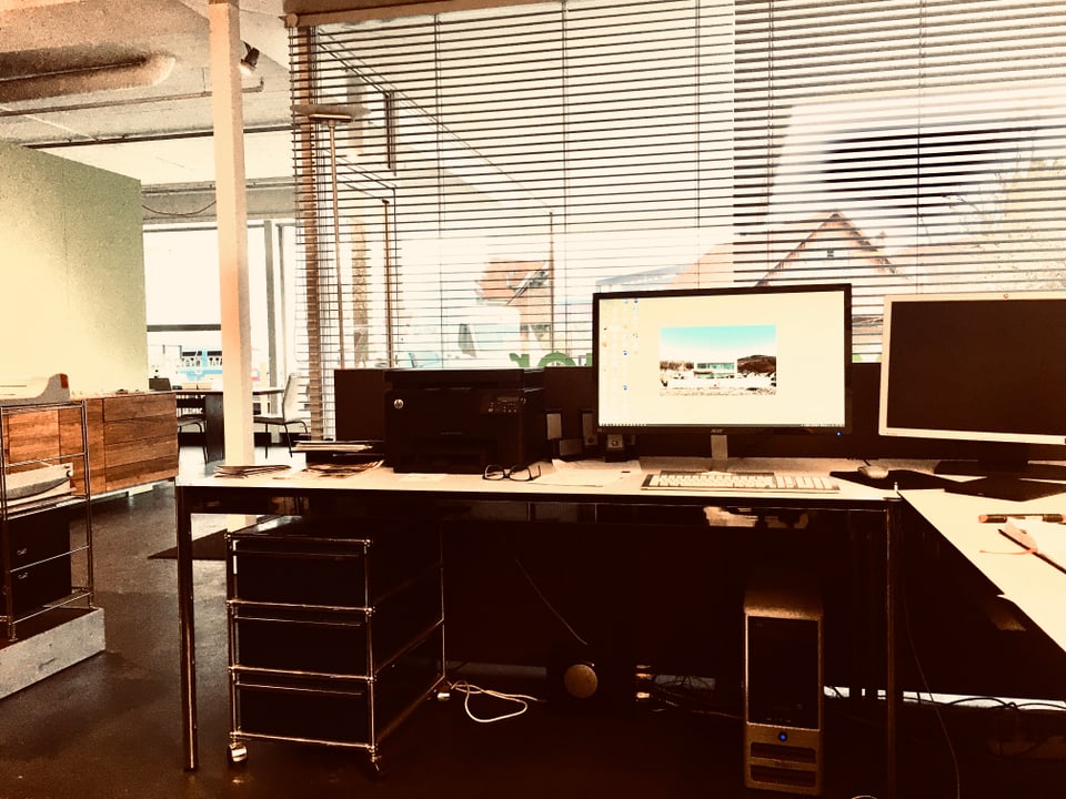 Ein Schreibtisch mit zwei Bildschirmen, dahinter ein geschlossener Rolladen