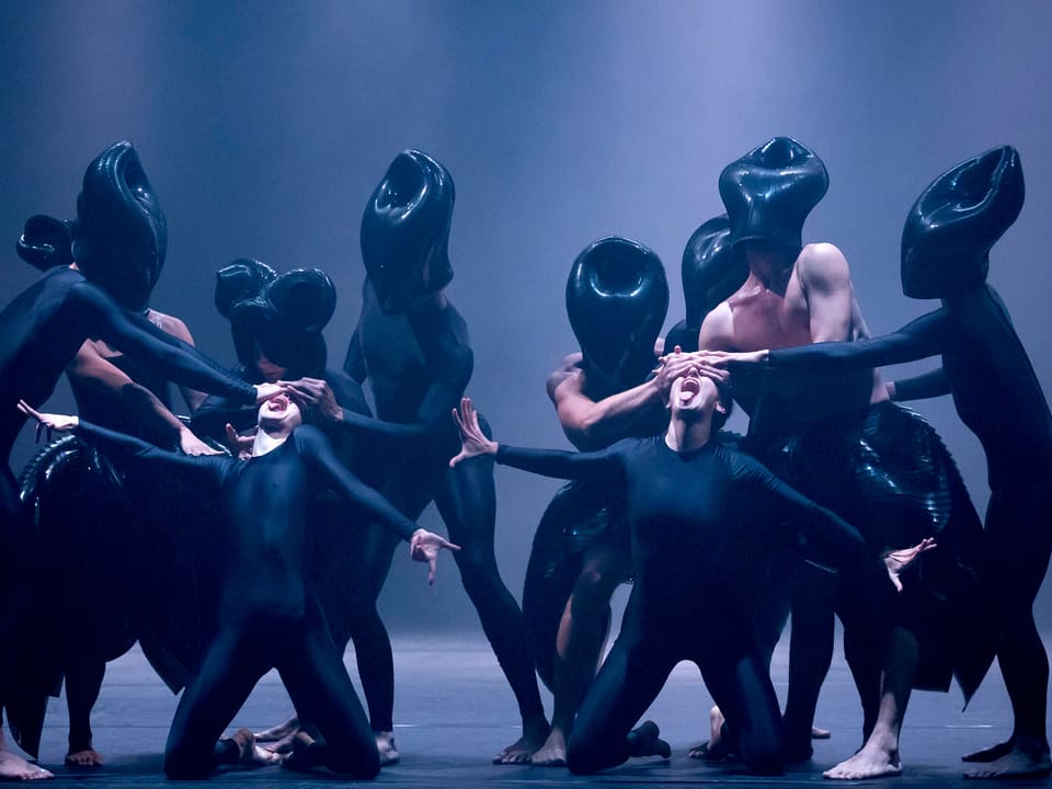 Eine Gruppe Tänzer mit schwarzen Masken hält zwei Menschen die Augen zu.