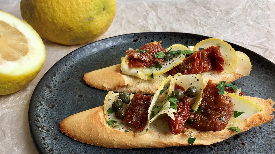 2 Bruschette mit fein geschnittenen Zedraten, eingelegten Tomaten, Kapern und Oliven auf einem Teller.
