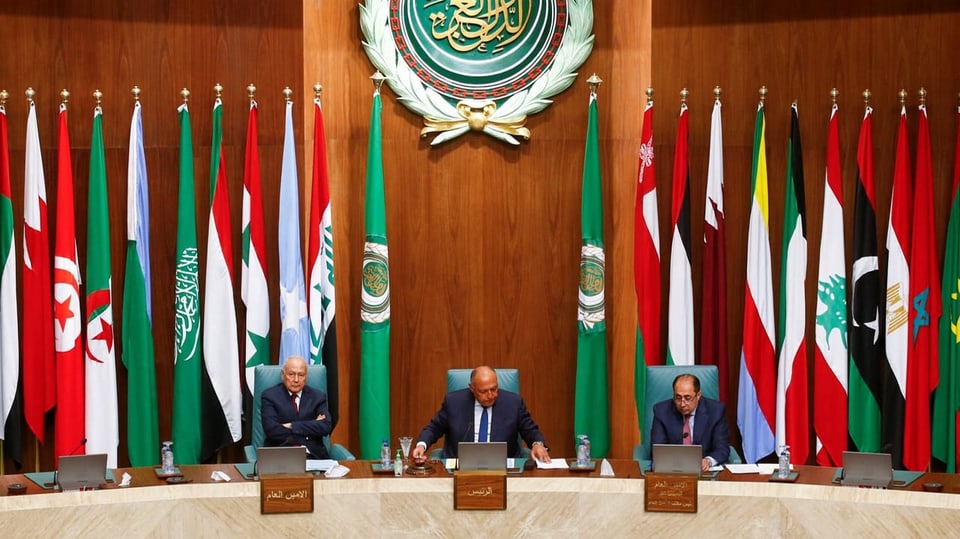Blick auf den Platz des Generalsekretärs der Arabischen Liga im Saal. In Hintergrund mehrere Flaggen und ein Emblem.