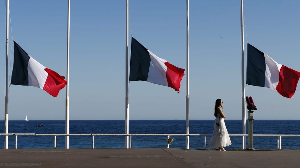 Drei französische Flaggen an der Promenade des Anglais auf Halbmast. Eine Frau mit Sonnenbrille läuft alleine.