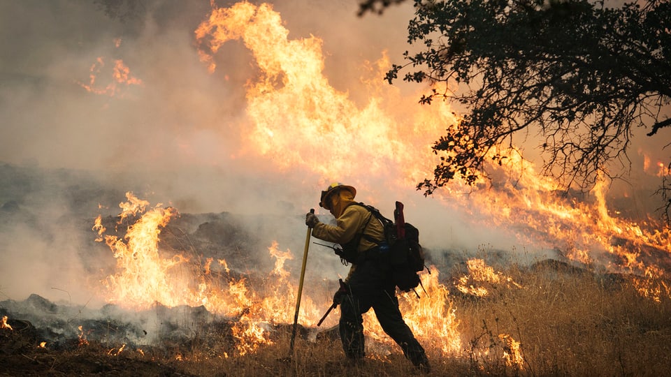 Ein Feuerwehrmann bekämpft Flammen am Boden, im Hintergrund lodern hohe Flammen