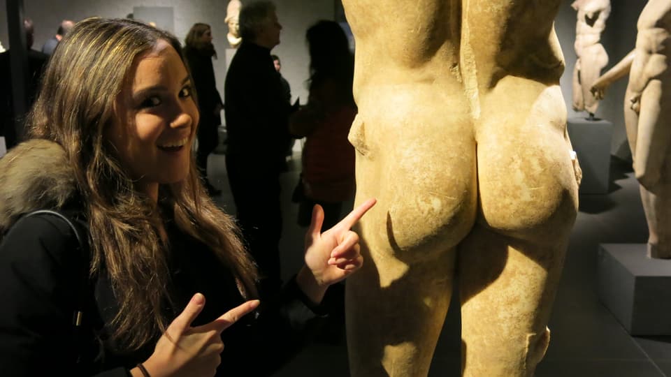 Junge Fraue Frau steht vor einer Skulptur und zeigt mit Finger auf den nackten Po der Skulptur.