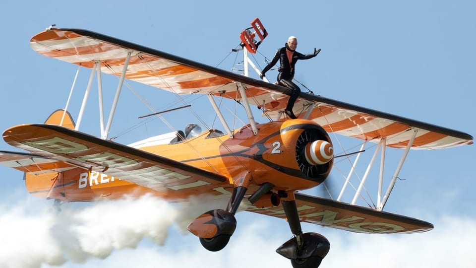 Ein oranger Doppeldecker fliegt durch die Luft. Ein Akrobat sitzt auf dem oberen Flügel. 