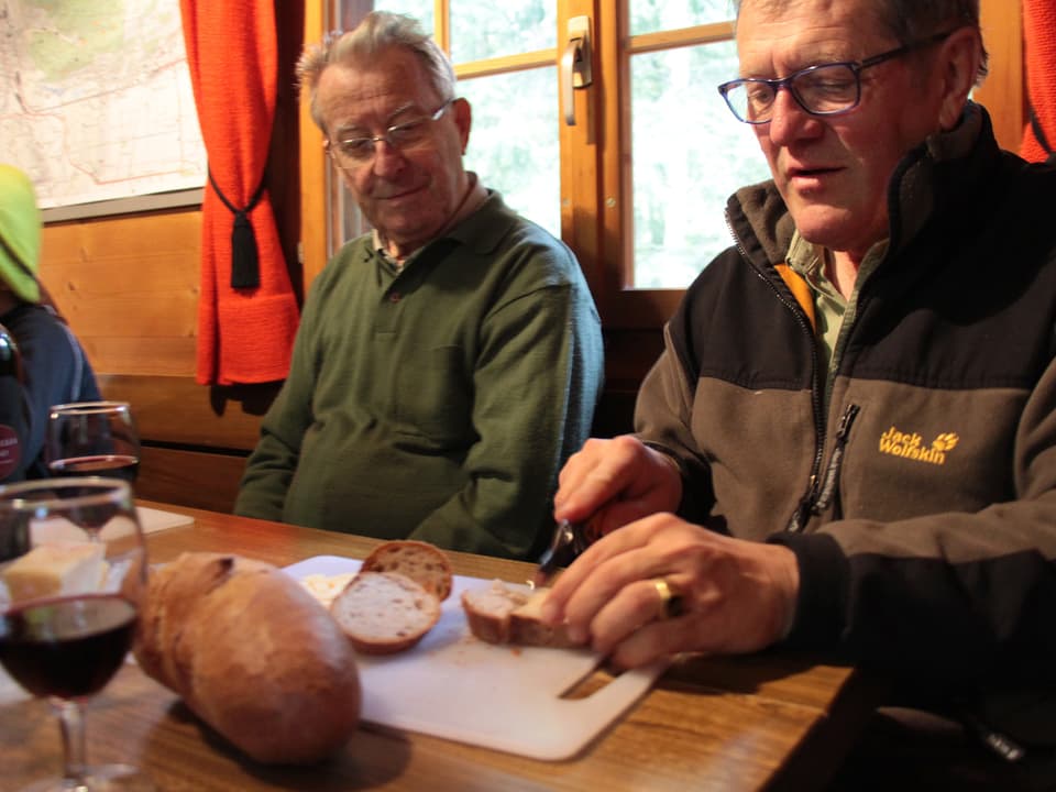 Zwei Männer sitzen in einer Hütte am Tisch und einer schneidet Brot. 