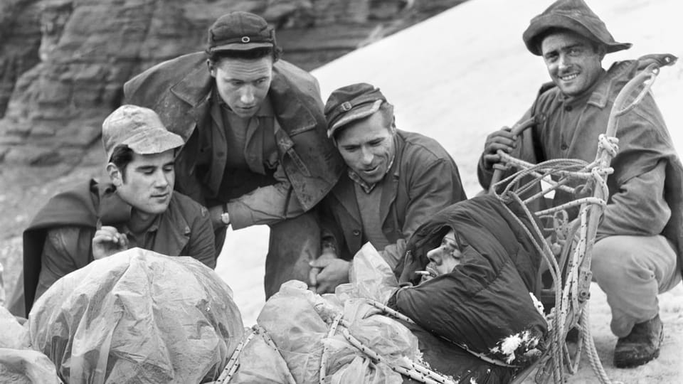 Schwarzweissfoto: Vier Männer knien neben einem Rettungsschlitten, auf dem ein weiterer, dick eingepackter Mann liegt. Einer der Männer schaut lächelnd in die Kamera